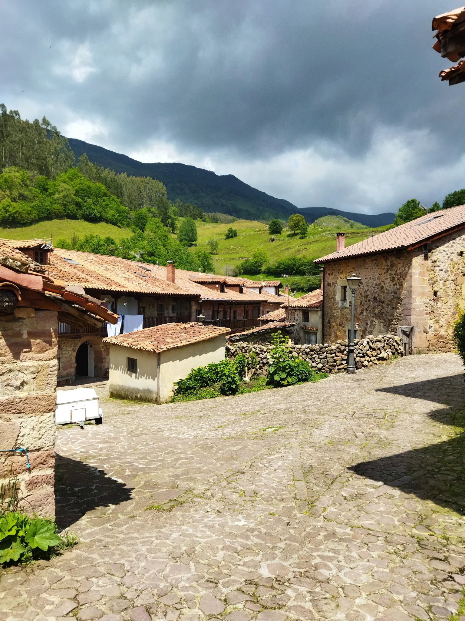 Conjunto de casas de piedra con un monte de color verde y árboles de fondo. El cielo está lleno de nubes. Parada obligatoria de la ruta en coche por Cantabria.