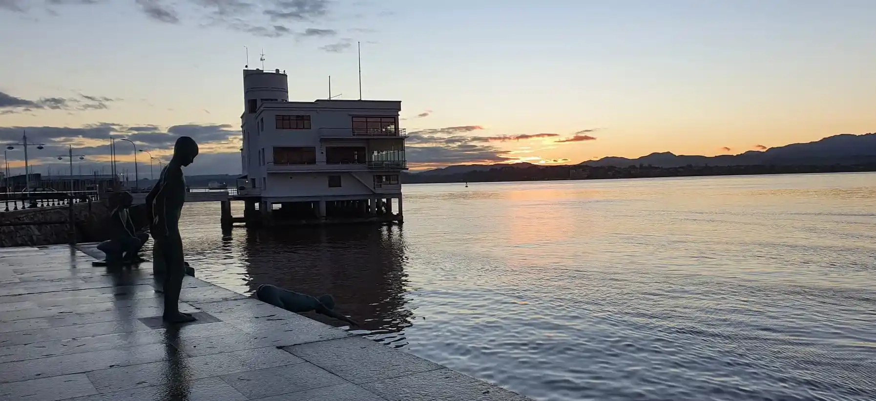 Foto de portada unas estatuas de unos niños tirándose al agua con un edificio al fondo dentro del agua de la bahía