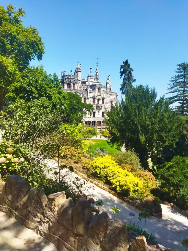 En el frente árboles y un camino de piedras. Detrás, un palacio de piedra en la Quinta da Regaleira.