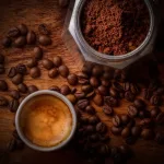Taza de café sobre madera, granos de café y cafetera italiana abierta con café molido