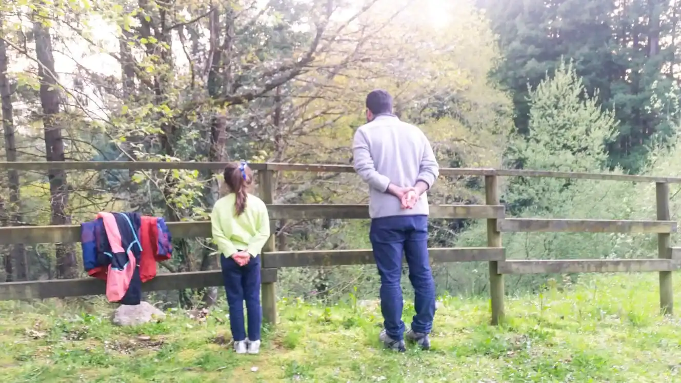 Una niña y un hombre con las manos detrás mirando por encima de una barandilla de madera con un bosque detrás. Bosque de las Secuoyas en Cabezón de la Sal.