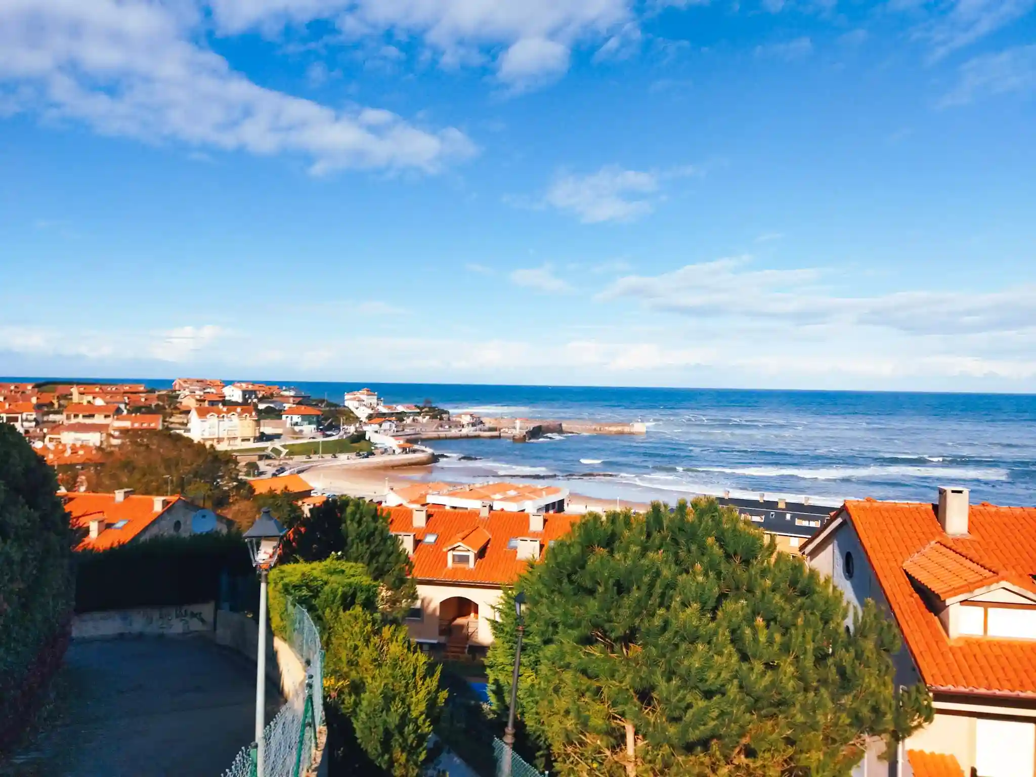 Vista desde un alto de una playa y diversas casa. Con el cielo completamente azul y algunas nubes sueltas. Comillas.