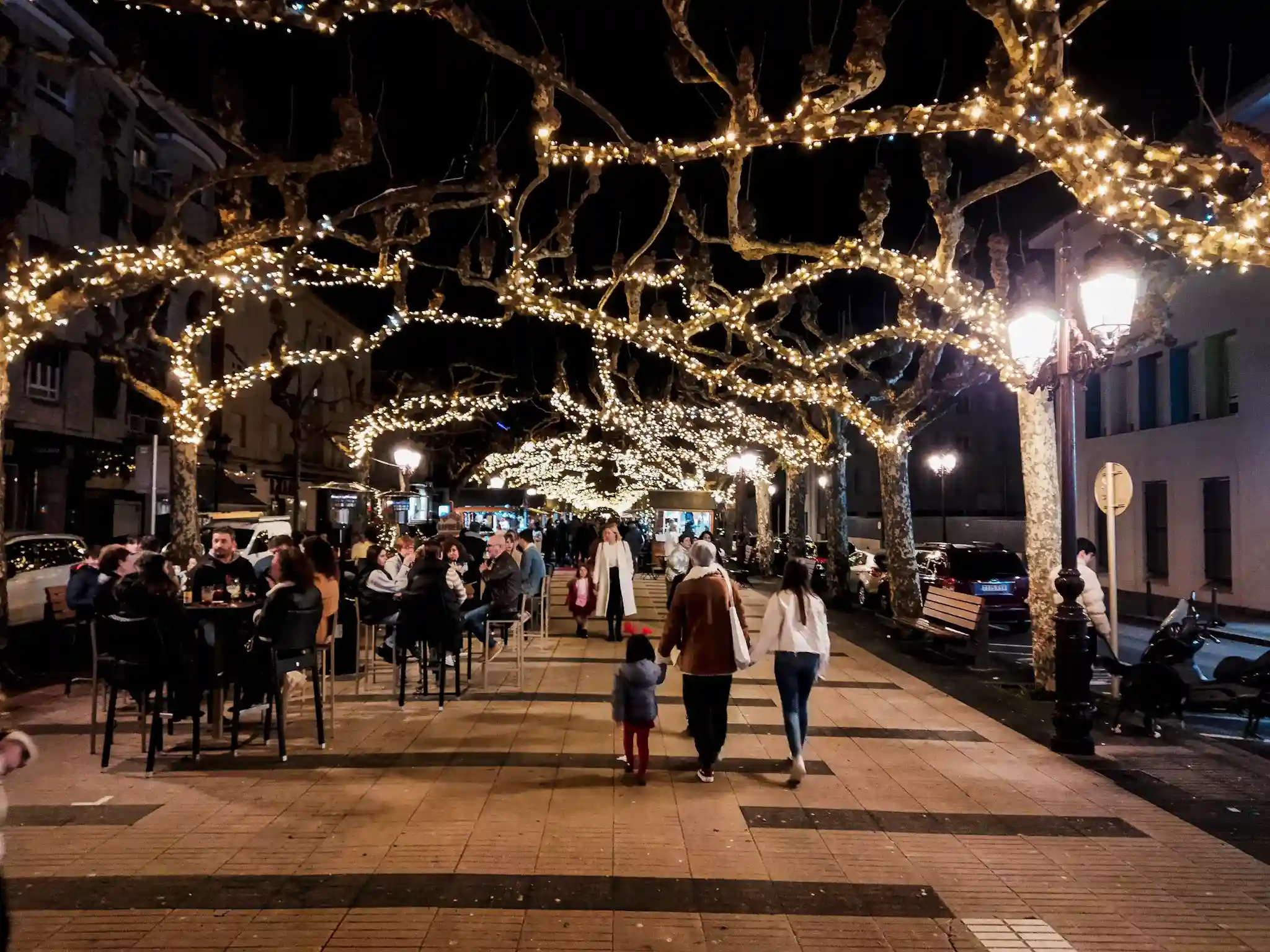 Tres personas caminando y gente sentada tomando algo. A los lados hay árboles y puestos navideños vendiendo.