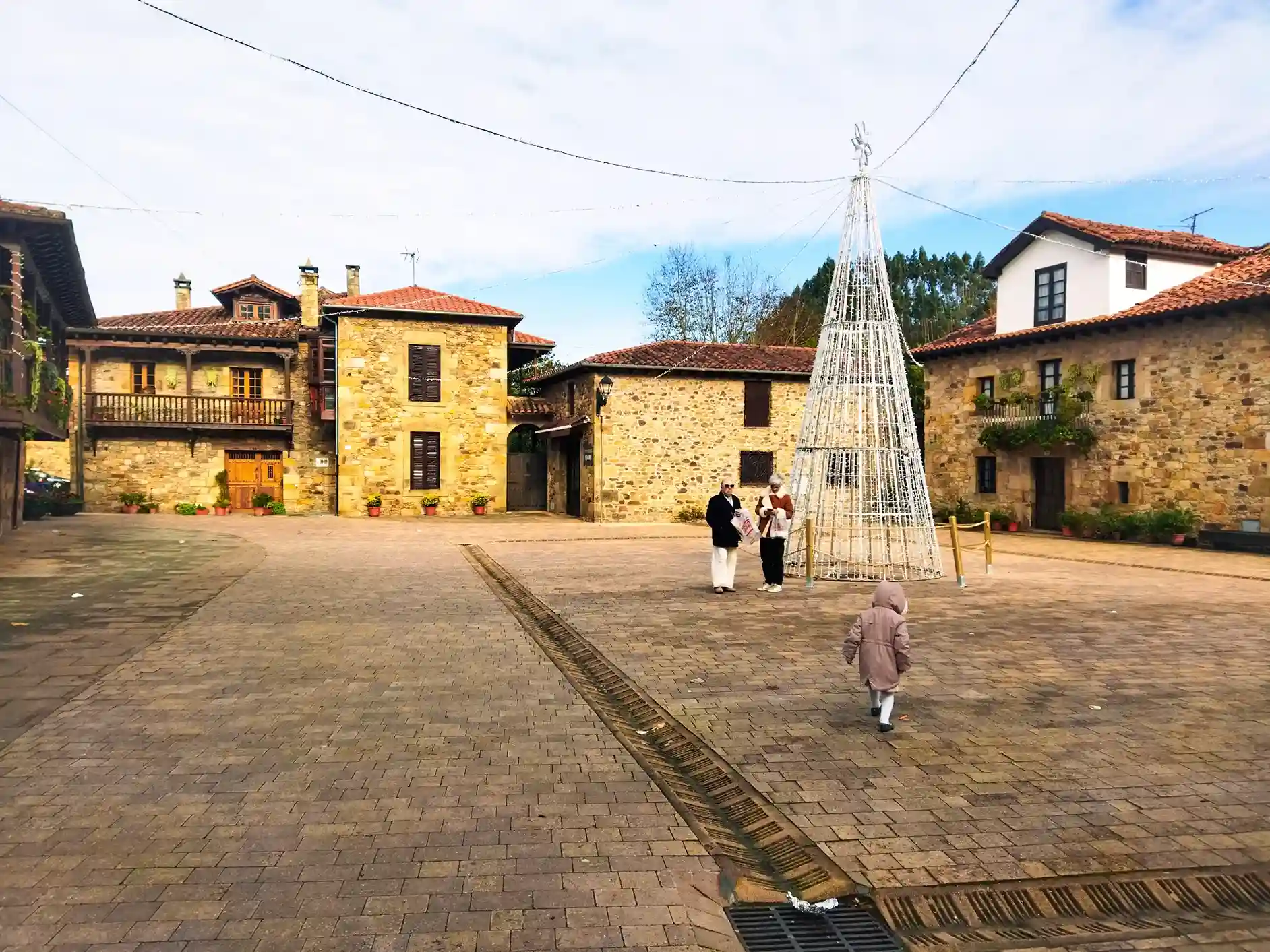 Plaza de suelo empedrado con casa de piedra alrededor y dos mujeres y una niña en medio