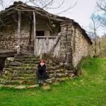 Cabaña pasiega con una mujer sentada las escaleras. Pisueña. Ruta circular en coche por los Valles Pasiegos.