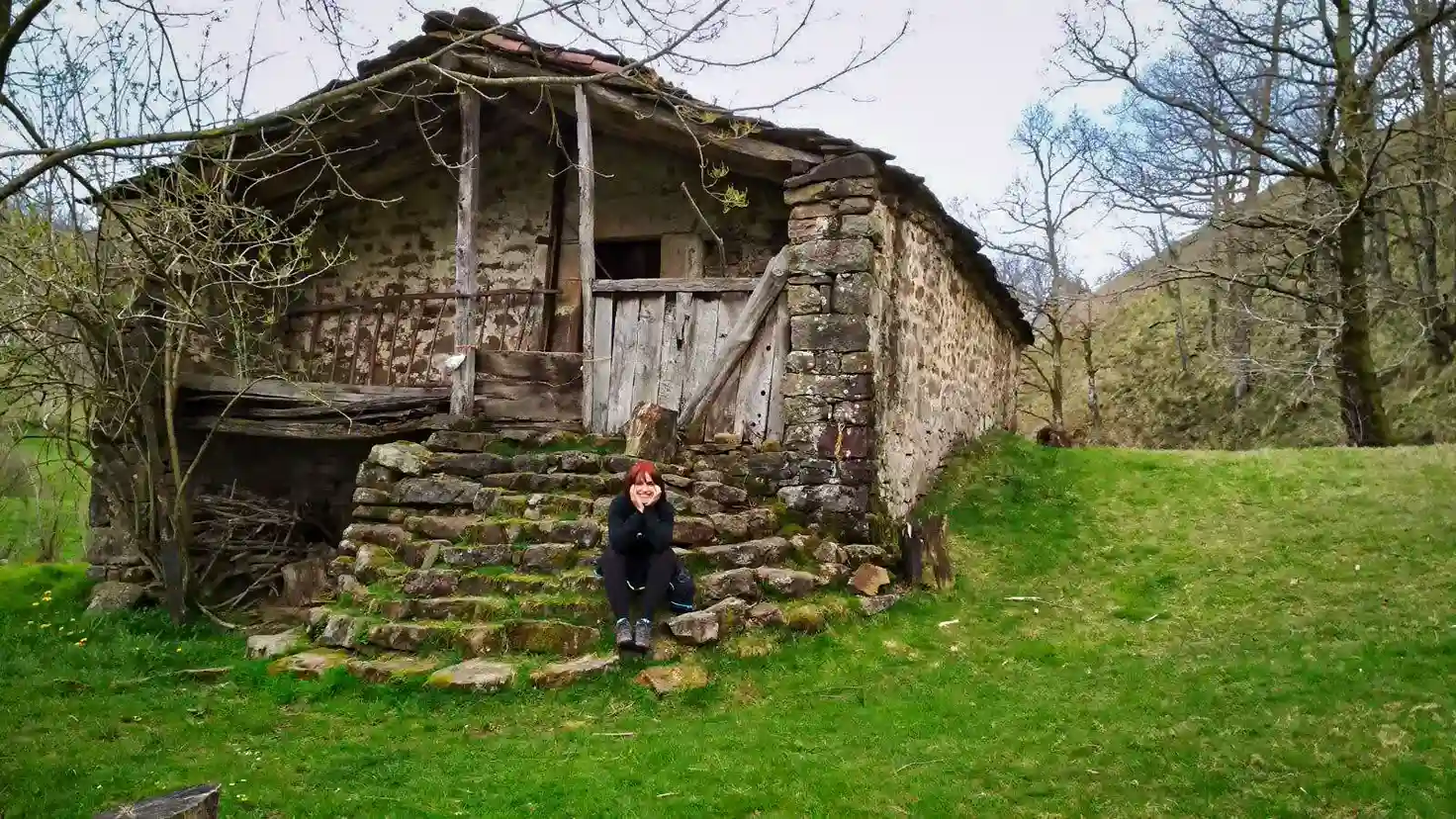 Cabaña pasiega con una mujer sentada las escaleras. Pisueña. Ruta circular en coche por los Valles Pasiegos.