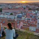 Two women sitting on the grass by a railing look out over the city of Lisbon under an orange sky from a viewpoint