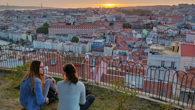 Dos mujeres sentadas en la hierba junto a una barandilla contemplan la ciudad de Lisboa bajo un cielo anaranjado desde un mirador